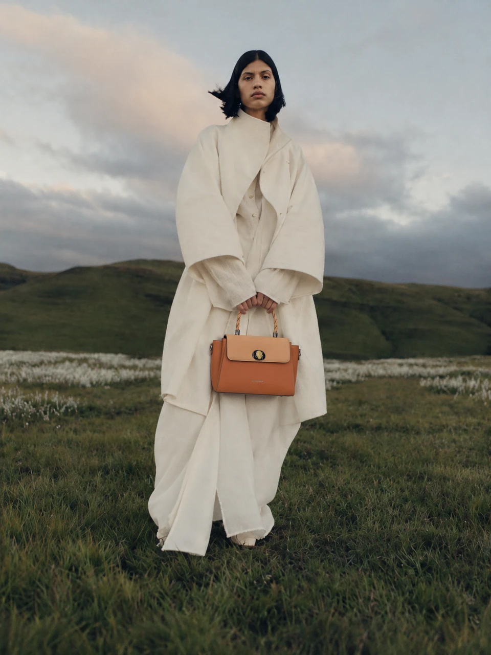 Model in an ivory coat holding the caramel and apricot SU QUEENA3234 bag in a windswept meadow.