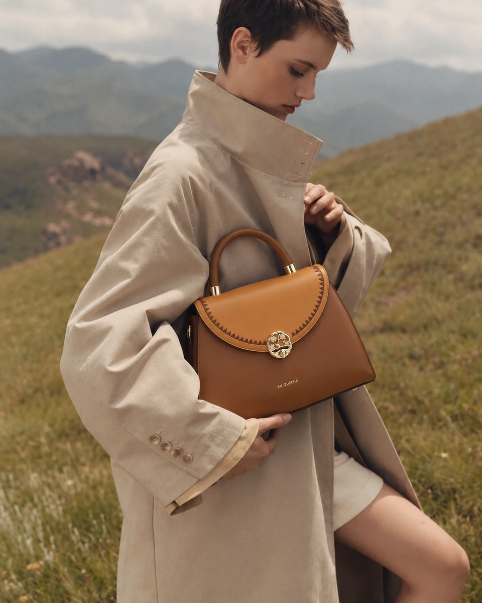 Model in a beige high-collar trench supporting the coffee and tan SU QUEENA 3131 bag against a hillside.