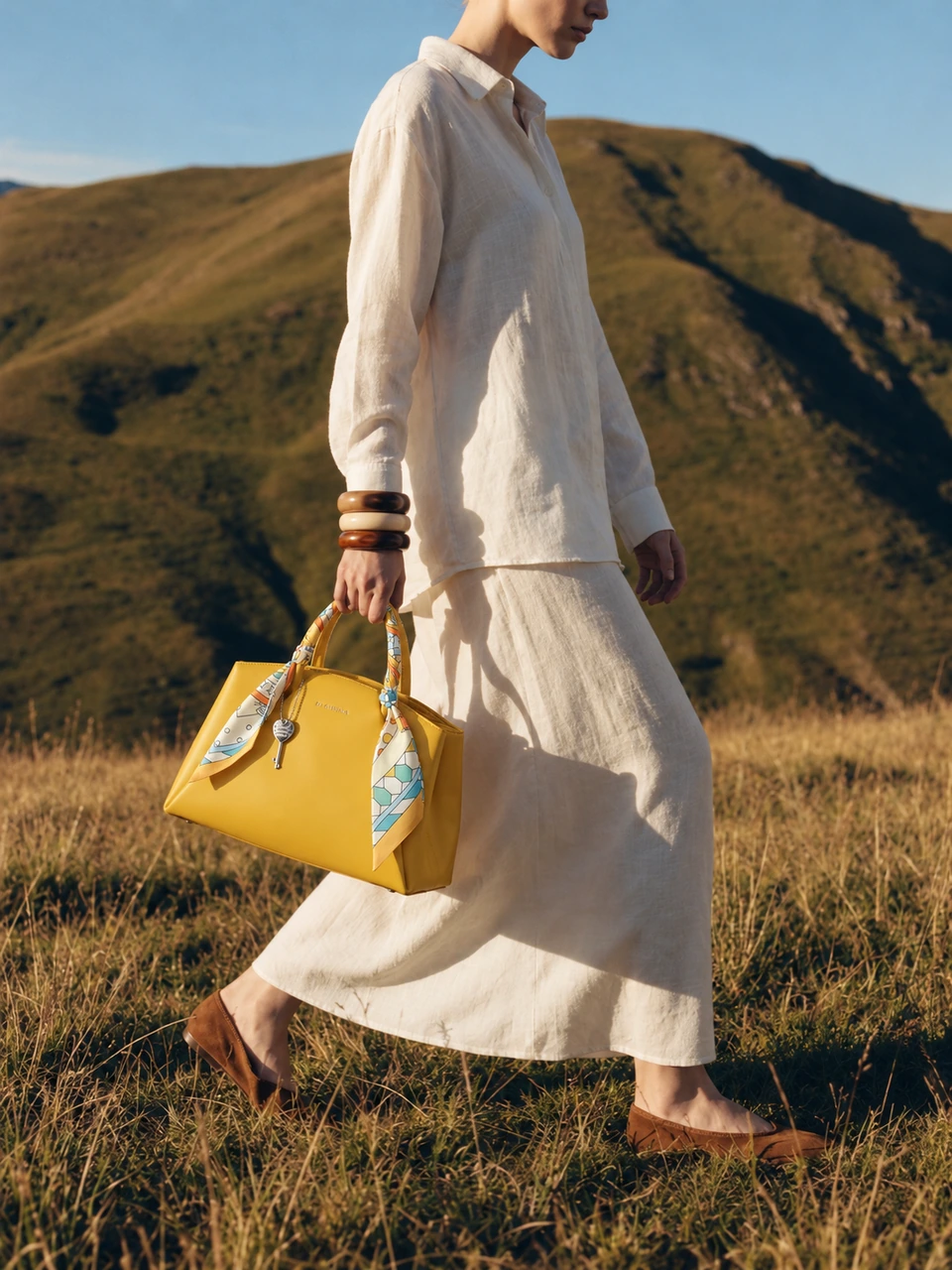 SU QUEENA yellow scarf-handle bag carried with ivory linen in sunlit hills.